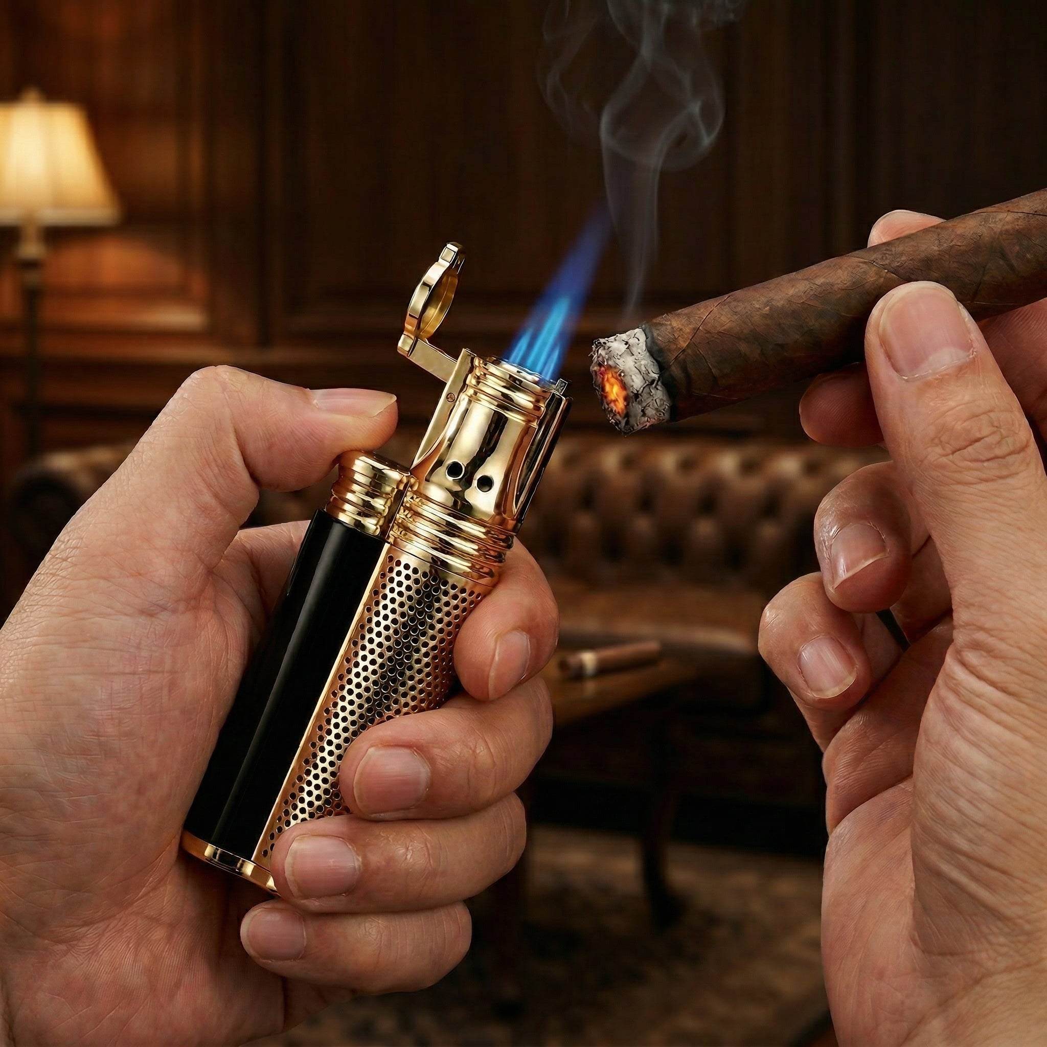 Lifestyle product photograph showing a person's hands using a gold and black triple-jet torch lighter to light a premium cigar. A bright blue flame meets the glowing cigar tip in a dimly lit, high-end lounge setting with a leather Chesterfield sofa in the background.