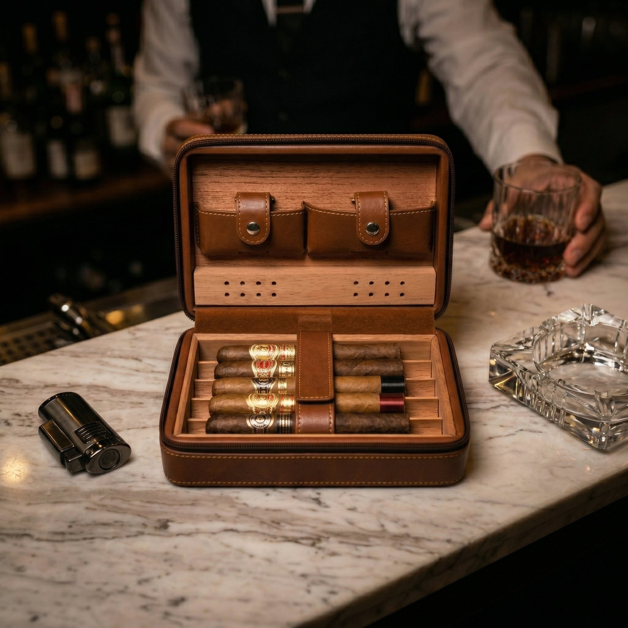 Luxury lifestyle view of an open brown leather travel cigar humidor on a marble bar top. Features a Spanish cedar wood interior with five premium cigars, integrated accessory pockets, and a nearby torch lighter and crystal ashtray, set against a sophisticated lounge background.