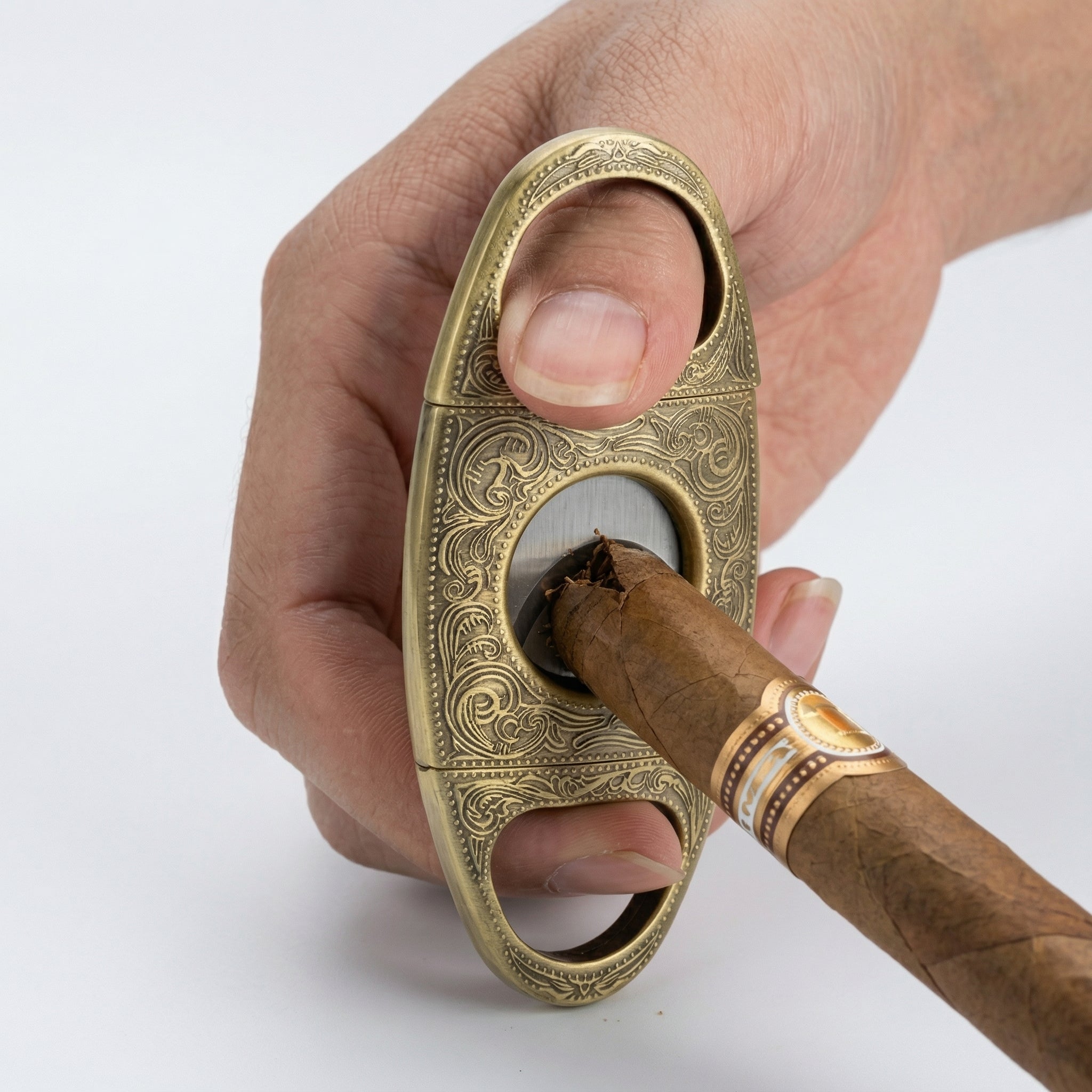 Action shot of a person's hand using the ornate antique brass double-guillotine cutter to slice the cap of a premium cigar. The image captures the moment the stainless steel blades close on the tobacco, set against a clean white background.