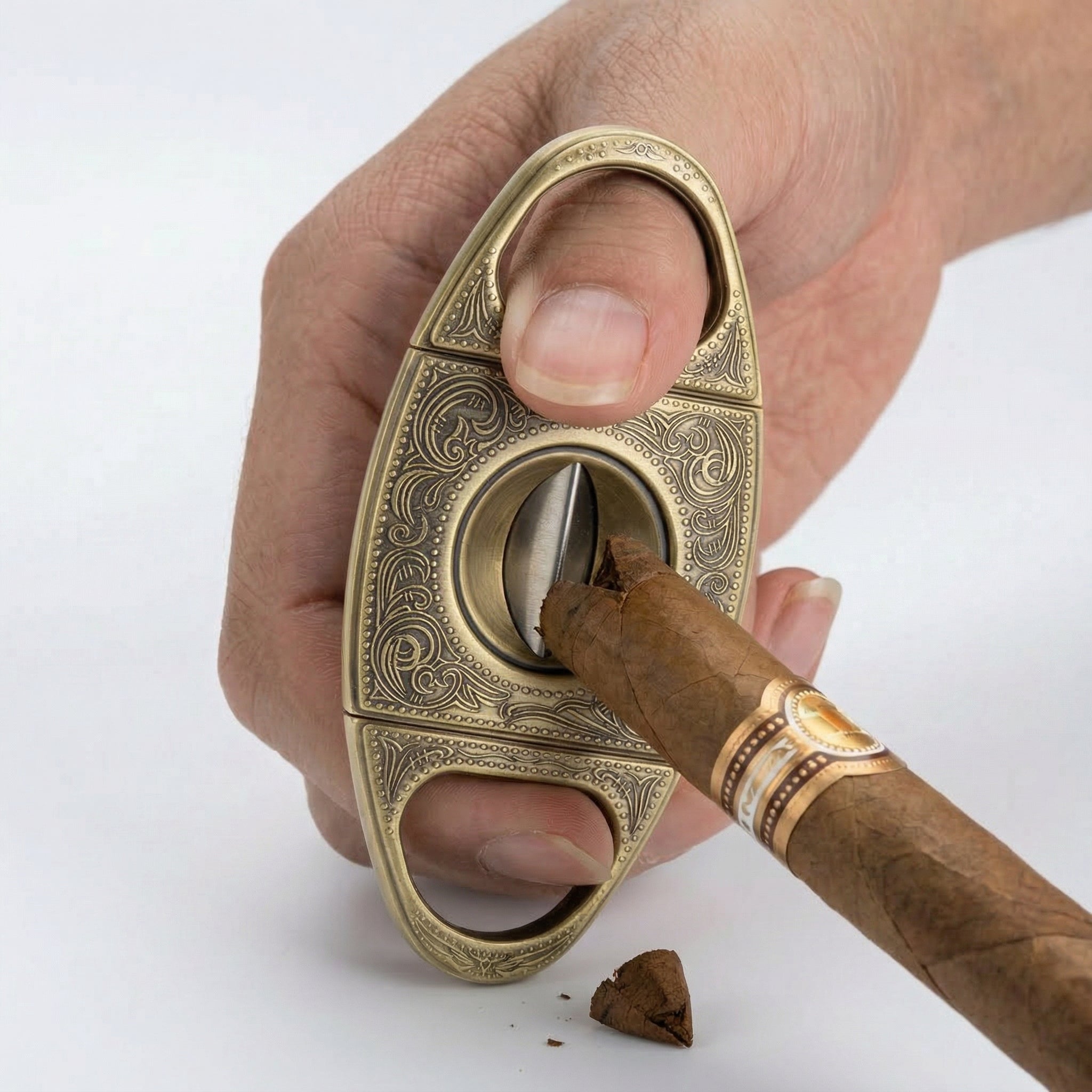 Action shot of a person's hand using an ornate antique brass V-cut cigar cutter. The image shows the device creating a precise wedge-shaped notch in the cigar cap, with the severed tobacco piece resting on a white background.
